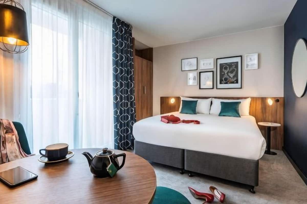 A hotel room with a bed, bedside table, lamp, and a view of the outdoors.