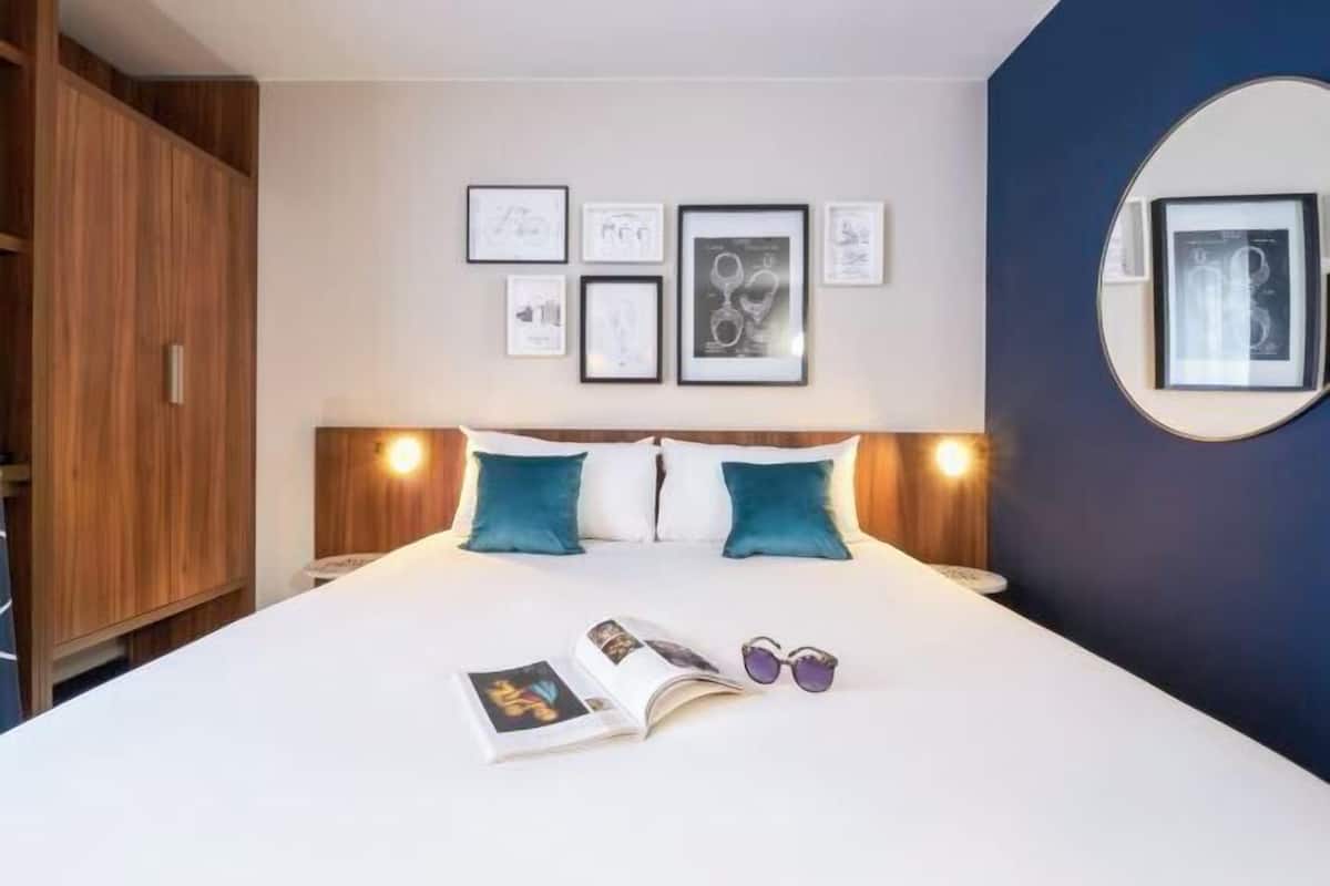 A neatly made bed with a book and sunglasses, a wall with framed pictures, and a round mirror.