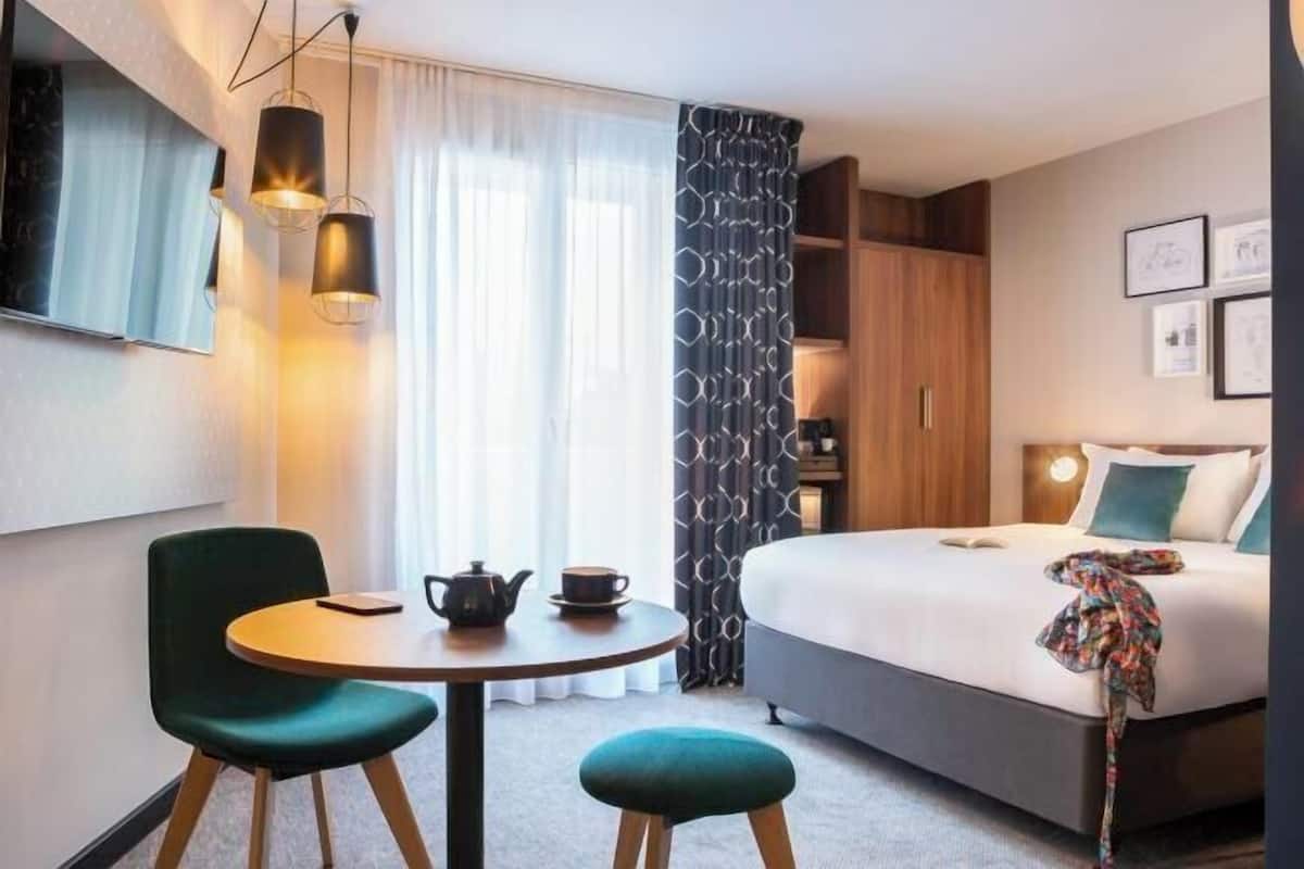 Une chambre d’hôtel avec un lit, une table ronde entourée de chaises, une petite table avec une théière et une tasse, et une grande fenêtre avec des rideaux.