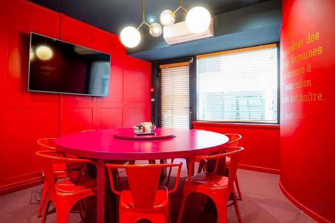 A red conference room with a round table, chairs, a flat-screen TV, and a window with blinds.