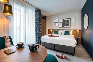 Habitación de hotel con una cama, una mesa redonda con tetera, una computadora portátil y una silla.