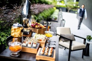 Una mesa de buffet con diversos alimentos y una jarra, ubicada al aire libre cerca de una silla y una mesita.
