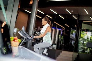 Een vrouw die een hometrainer gebruikt in een moderne sportschool met grote ramen en een mand met fruit.