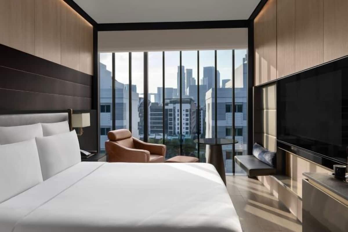 Una habitación de hotel moderna con una cama grande, una zona de estar con una silla y una mesita, un televisor y vistas a la ciudad a través de amplias ventanas.