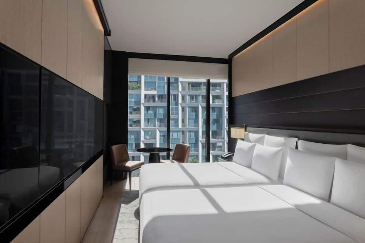 Habitación de hotel moderna con una cama grande, un escritorio y una silla. Se ve una ventana amplia con vistas a otros edificios.