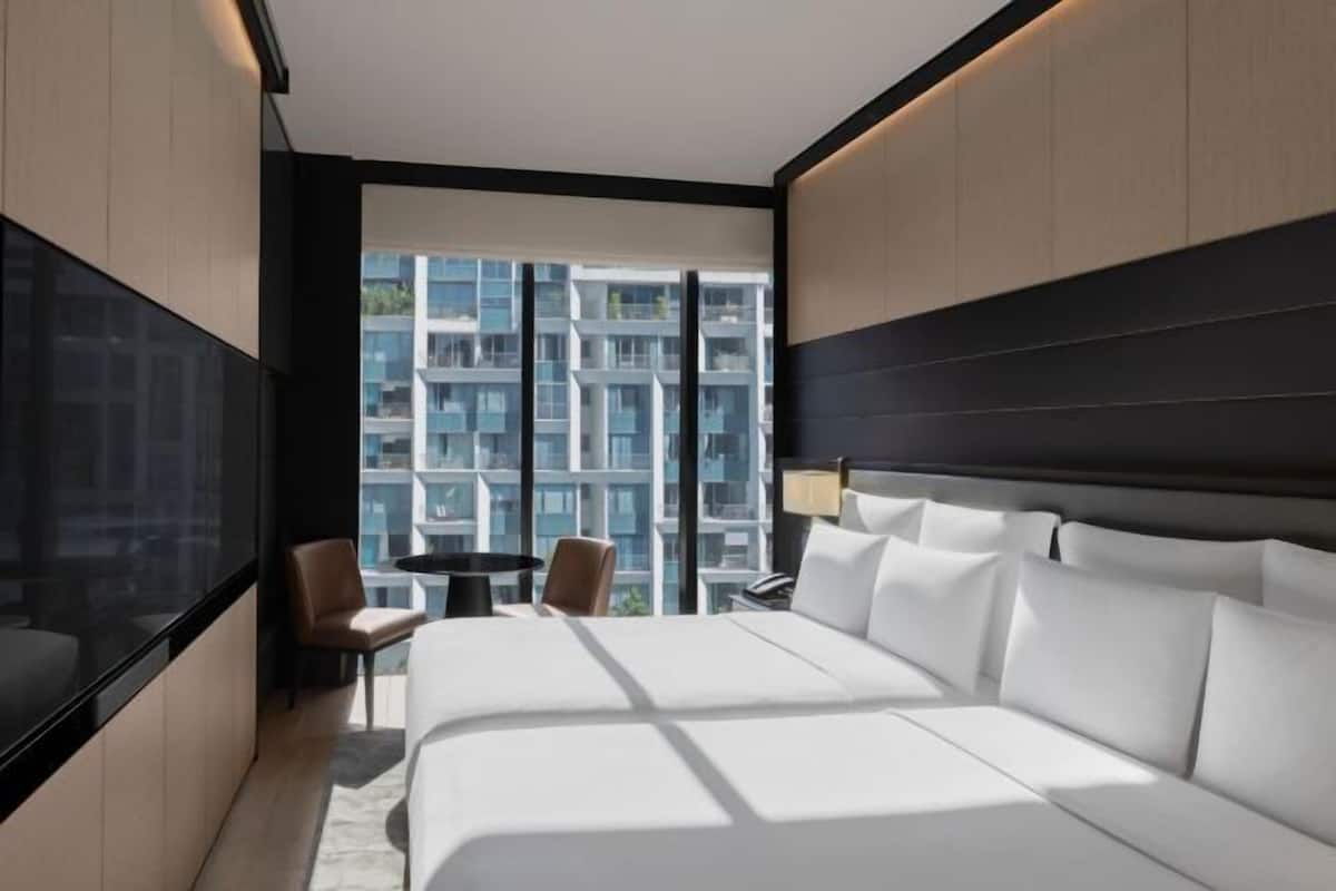 Habitación de hotel moderna con una cama grande, un escritorio y una silla. Se ve una ventana amplia que da a otras construcciones.