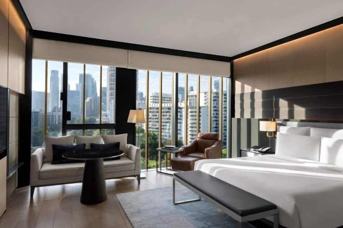 Una habitación de hotel moderna con una cama grande, un sofá, una silla y una mesita. La habitación tiene ventanas amplias con vistas a la ciudad.