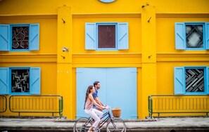 Een koppel fietst voorbij een geel gebouw met blauwe luiken en geometrische raamdecoraties.