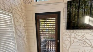 A room with a wooden floor, a black-framed door, a window with blinds, a ceiling light, and a wall-mounted painting. - Turquoise Triangle Tiny Homes (Cottonwood)