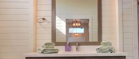 Ein Badezimmer mit einem Holzkosmetikschrank, einem großen Spiegel, einem Fenster über dem Spiegel und Blick auf Bäume.