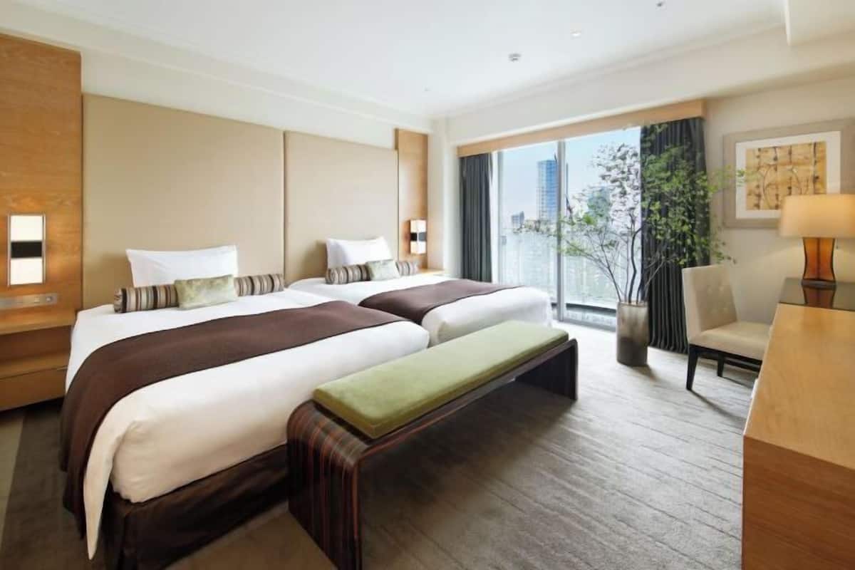 Habitación de hotel con dos camas, un banco, un escritorio, una silla, un ventanal con vistas y una planta.