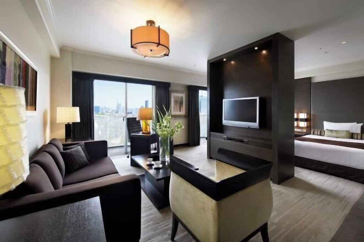 Habitación de hotel moderna con sofá, mesa de centro, televisor, cama y un amplio ventanal con vistas a la ciudad.