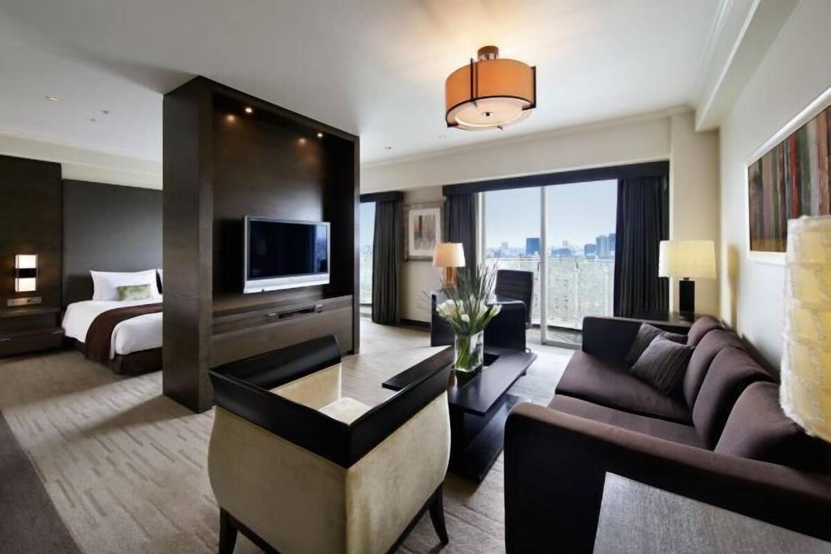 Habitación de hotel moderna con una cama grande, televisor, escritorio y sofá. Cuenta con un ventanal amplio con vistas a la ciudad.