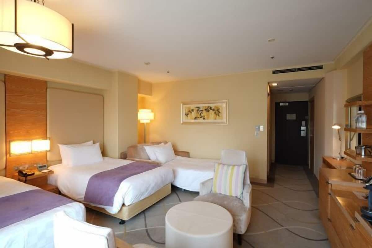 Habitación de hotel con dos camas, una silla, un otoman redonda, un cuadro en la pared y un escritorio con una lámpara.