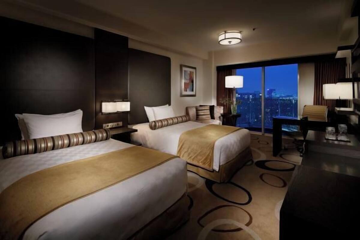 Habitación de hotel con dos camas, un escritorio, una silla, un ventanal grande y vistas a la ciudad, y una lámpara.