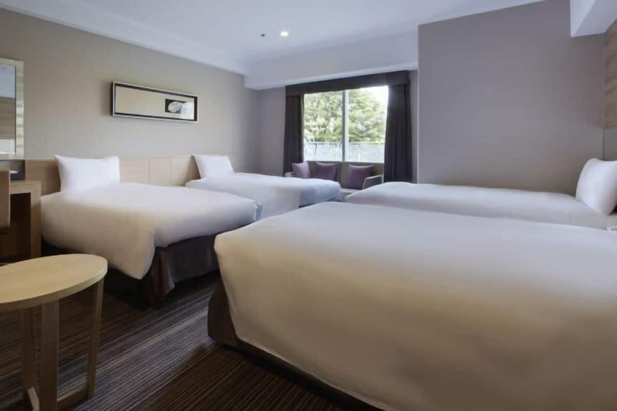 Una habitación de hotel con tres camas, una mesita redonda pequeña, un ventanal grande con cortinas y un cuadro en la pared.