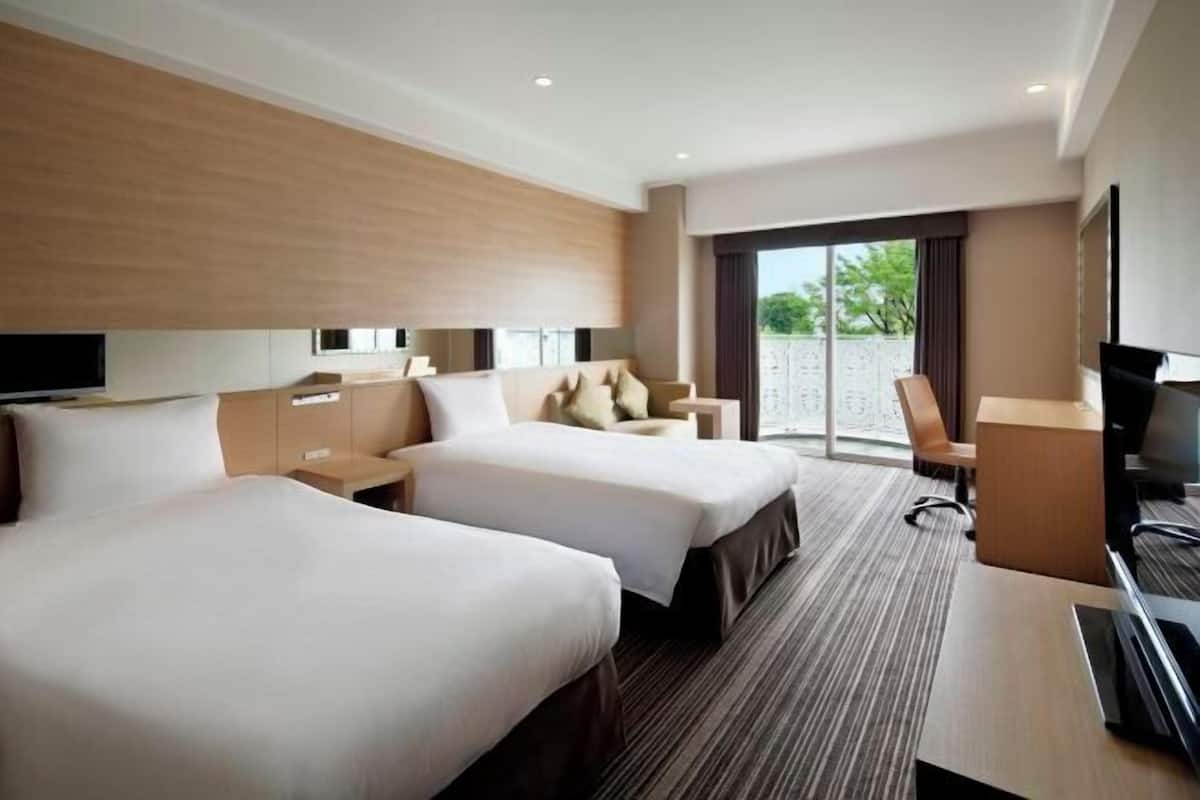 Habitación de hotel con dos camas, un escritorio, televisor y un amplio ventanal con vistas al exterior.