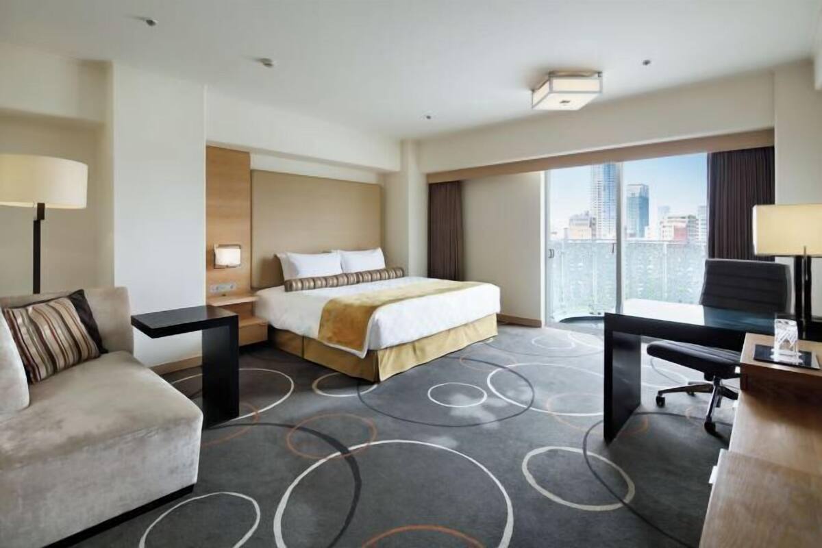 Habitación de hotel con cama, escritorio, sillón, sofá y un amplio ventanal con vista a la ciudad.