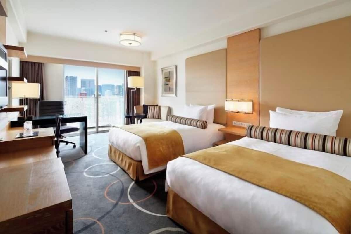 Habitación de hotel con dos camas, un escritorio, una silla y un ventanal con vista a la ciudad.