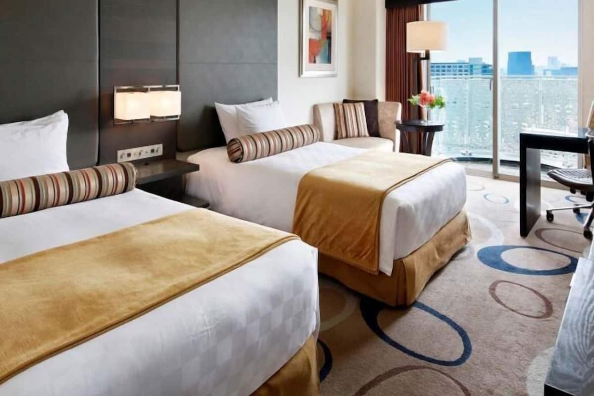 Habitación de hotel con dos camas, un escritorio, una silla, una lámpara y vistas a la ciudad.