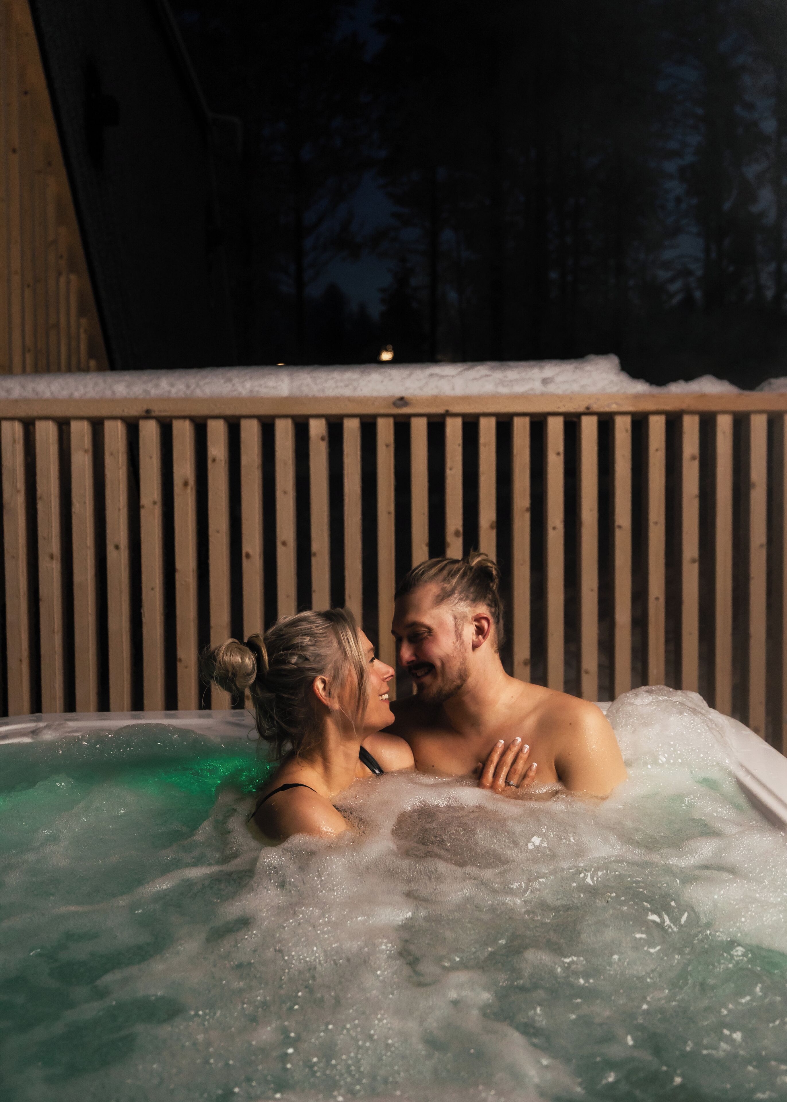 A couple in a hot tub with a wooden cabin background.