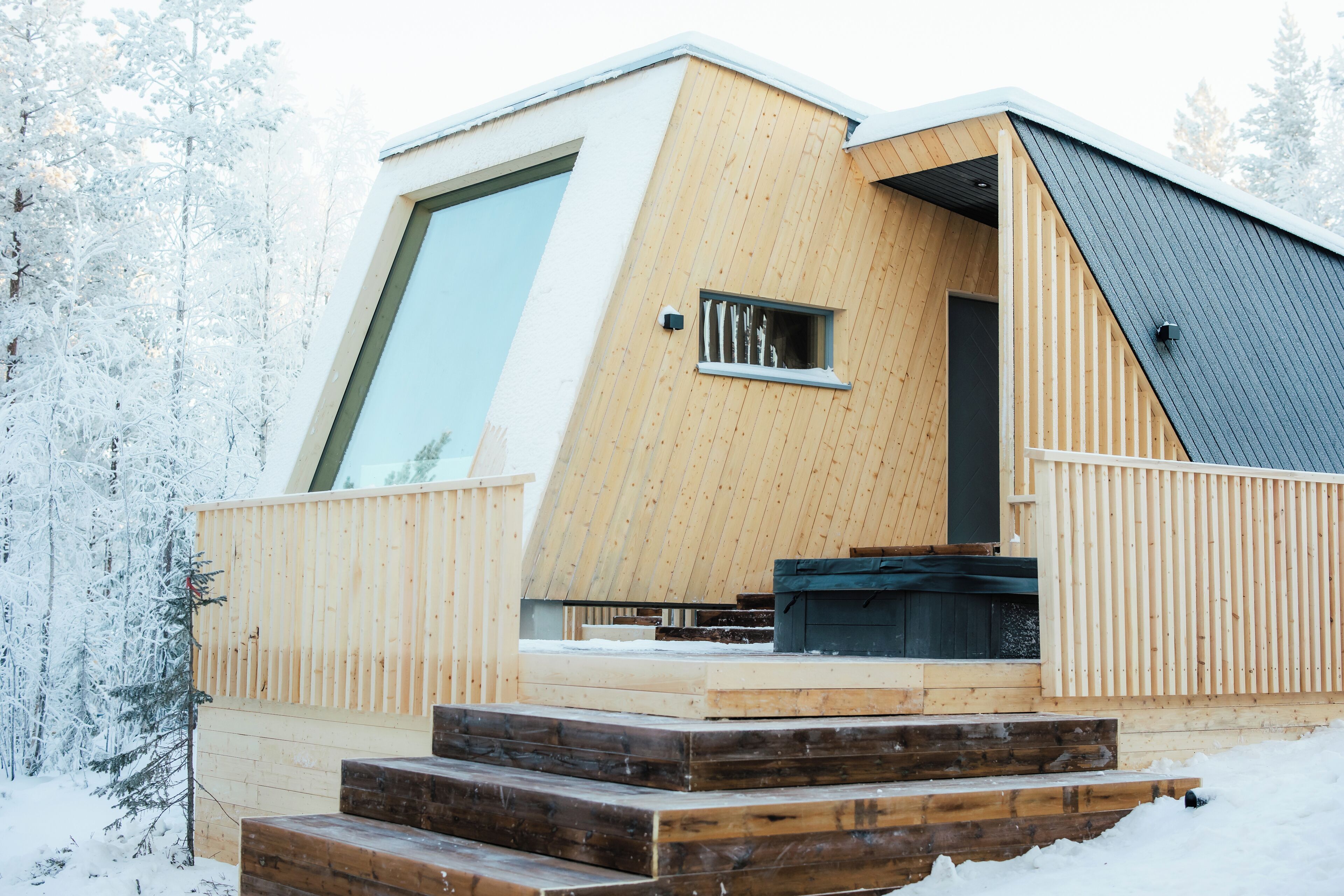 Una cabaña moderna de madera con un amplio ventanal, terraza de madera y un pequeño espacio de estar al aire libre.