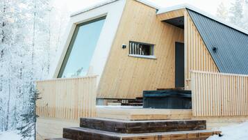 Una cabaña moderna de madera con un amplio ventanal, terraza de madera y un pequeño espacio de estar al aire libre.