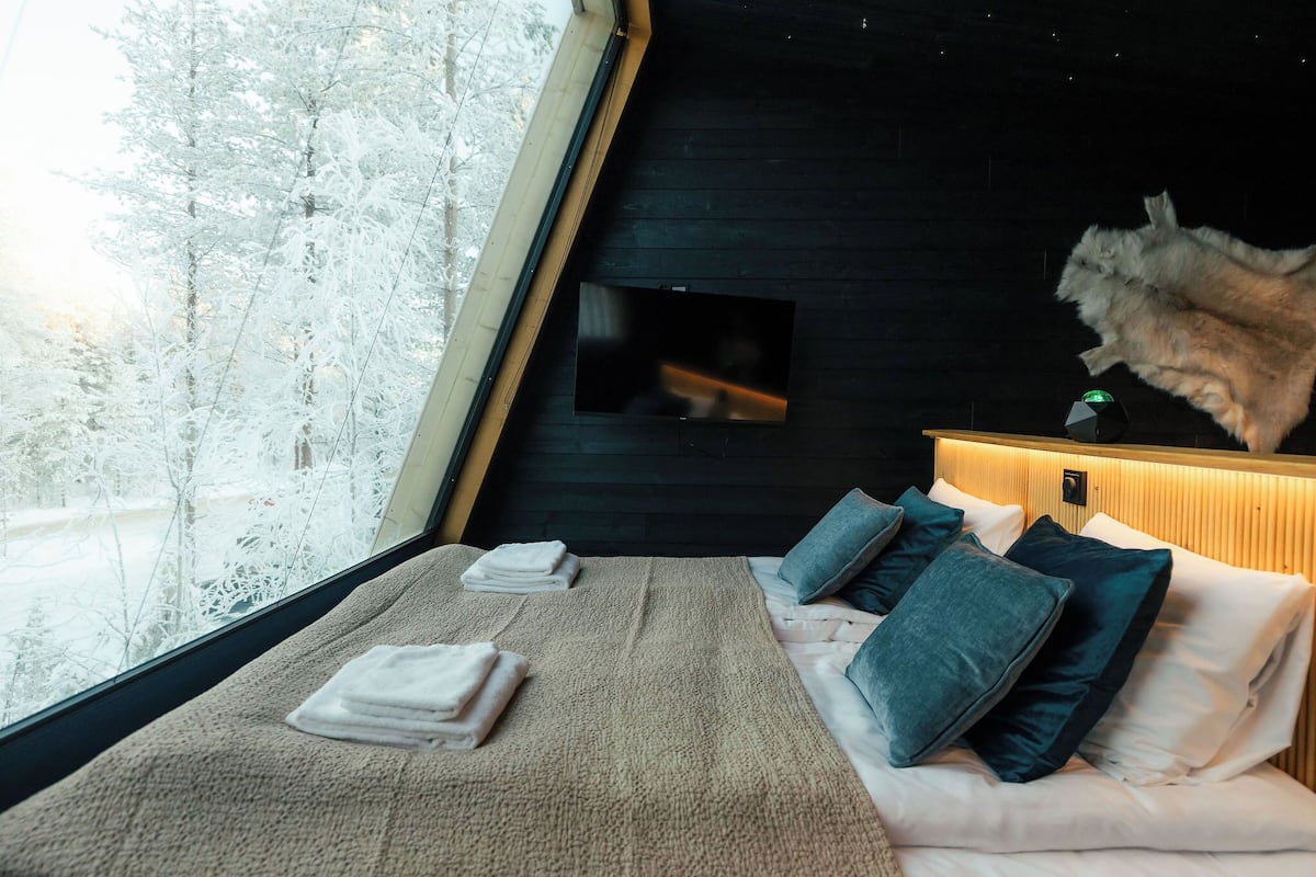 Een slaapkamer met een groot raam, een bed met kussens, een houten hoofdbord en een bontjas hangend aan de muur.