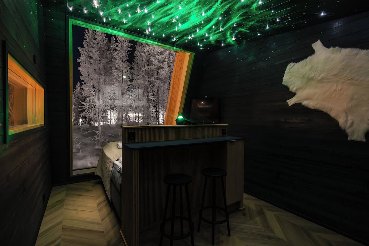 Een kamer met houten inrichting, een bed met een wit hoofdeinde, een bar met krukjes en een groot raam dat een besneeuwd bos toont.