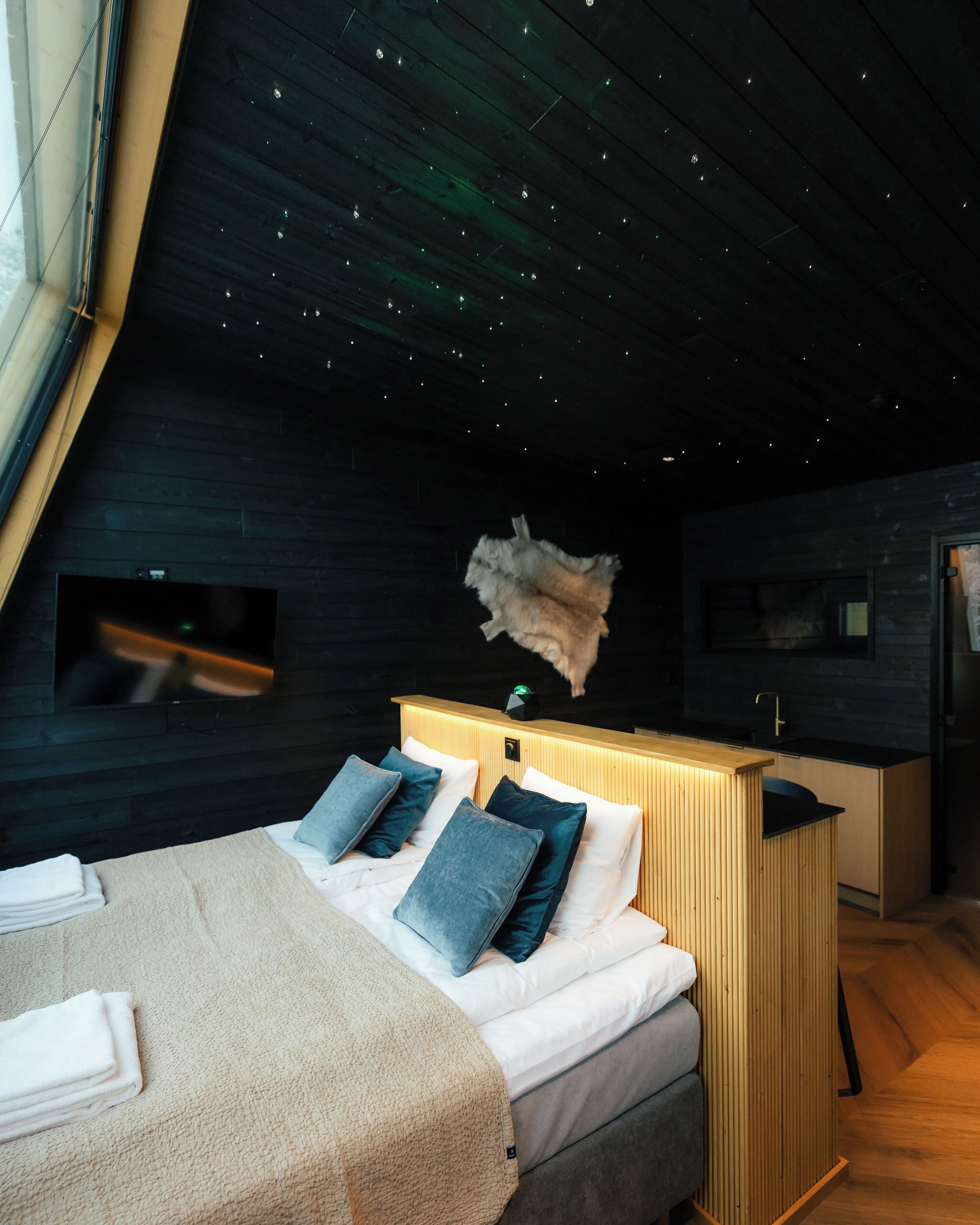 A modern bedroom with a sloped ceiling, a bed with white linens and blue pillows, a wooden headboard, and a small kitchen area.