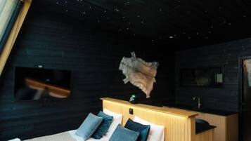A modern bedroom with a sloped ceiling, a bed with white linens and blue pillows, a wooden headboard, and a small kitchen area.