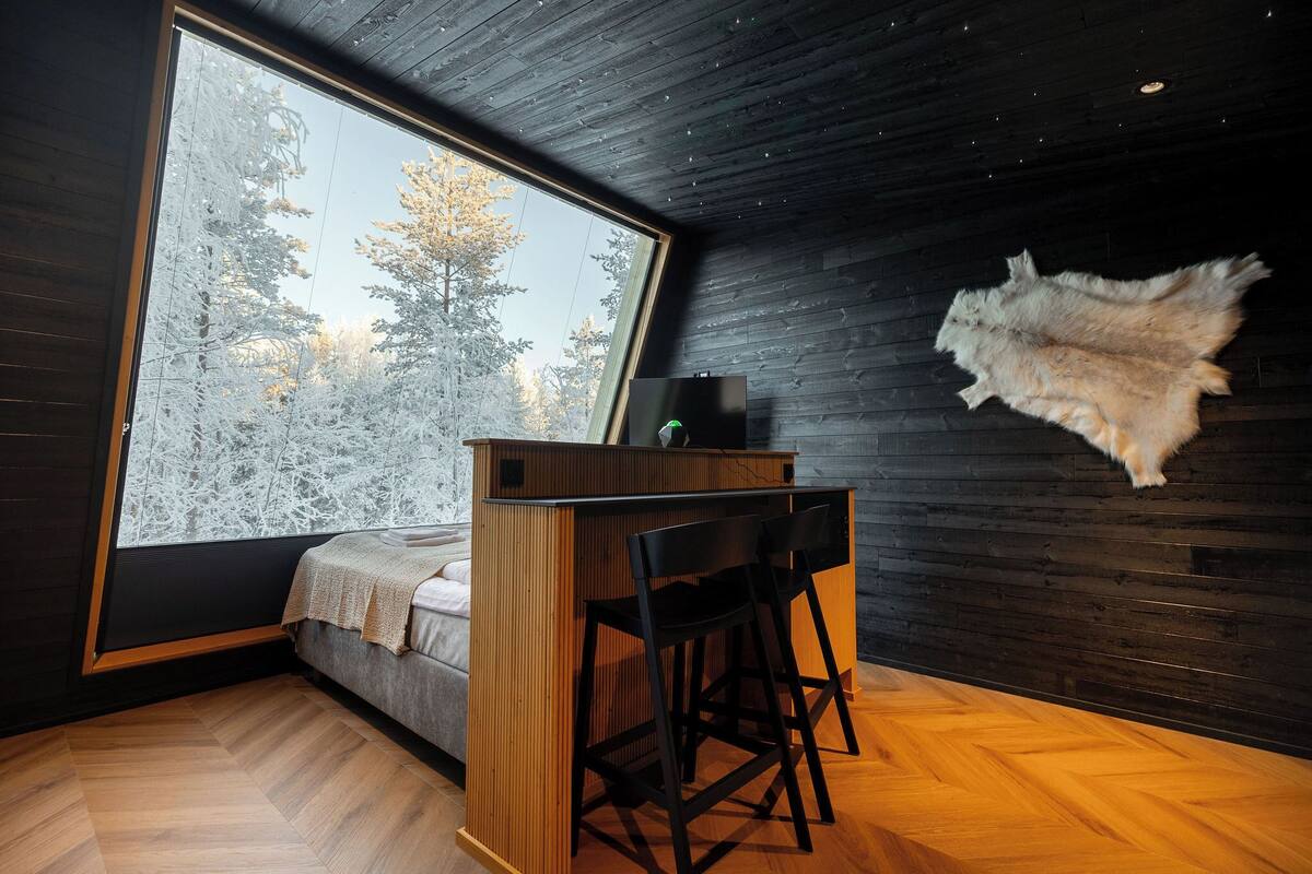 A modern room with a large window, a wooden desk, two black chairs, a bed, and a fur rug on the wall.