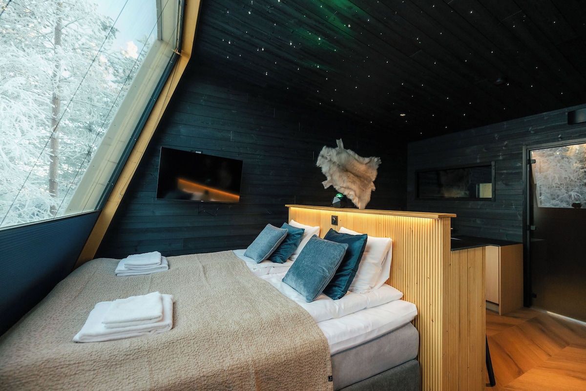 Une chambre à coucher moderne avec un lit, des oreillers, une tête de lit en bois, une petite télévision et une fenêtre donnant sur des arbres.