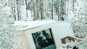 A snow-covered cabin with a large window, surrounded by trees. - Lapland Glow Chalets (Rovaniemi)