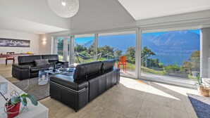 A modern living room with a black leather sofa, a glass coffee table, and a large window offering a scenic view of mountains and a lake. - Villa Mondacce With Jacuzzi & Lake Views (Minusio)