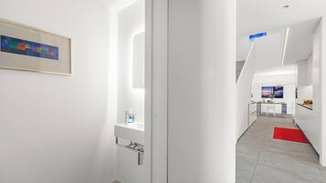 A modern hotel corridor with a minimalist design, featuring a white wall, a framed abstract painting, a small sink with a soap dispenser, and a red rug on the floor.