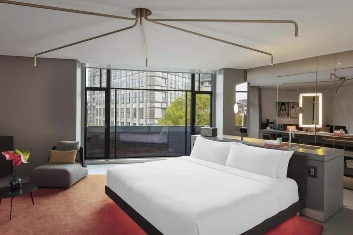 A modern hotel room with a large bed, a desk, a mirror, and a view of buildings through large windows.