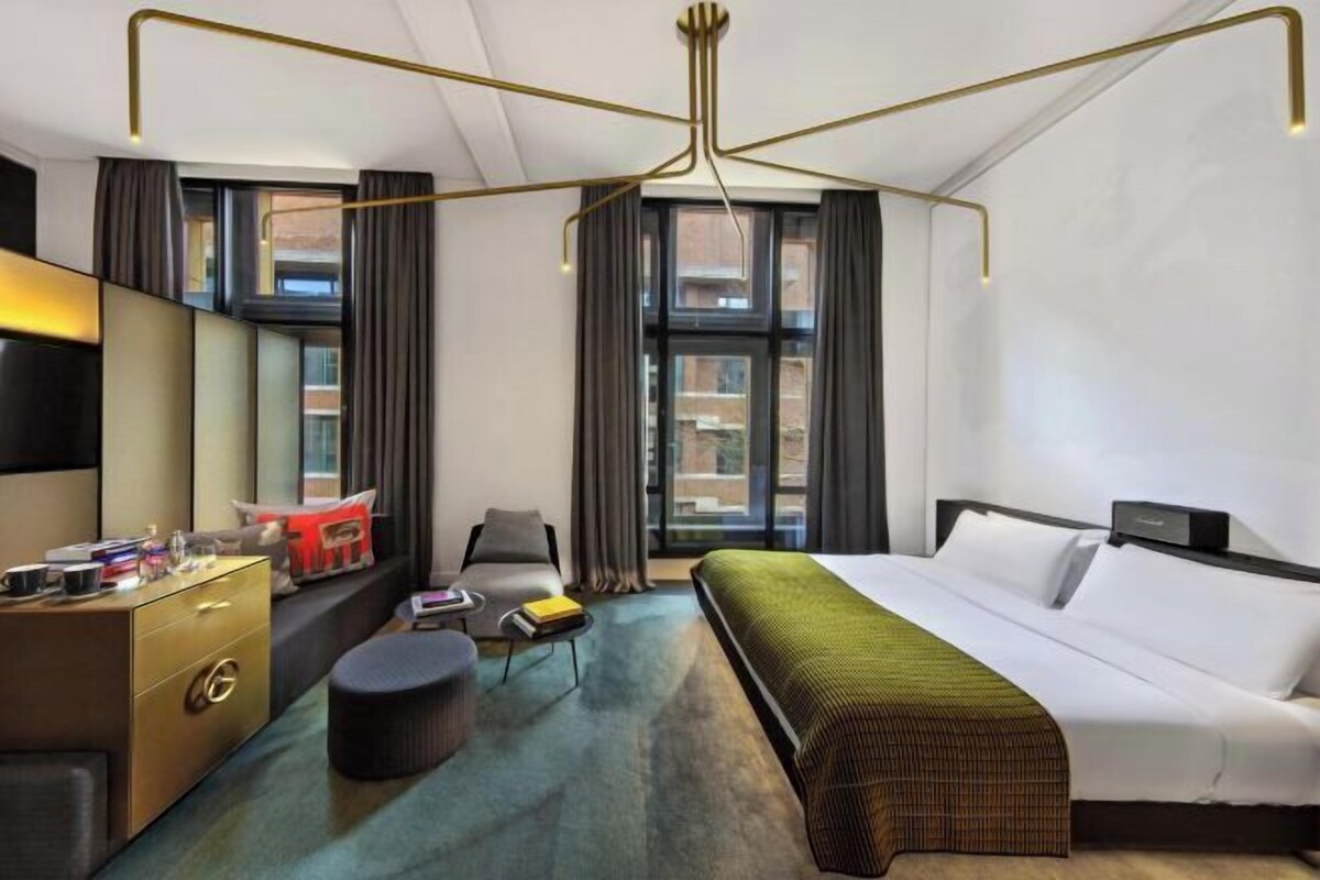 A modern hotel room with a large bed, a desk with a chair, a sofa, and a large window with curtains.