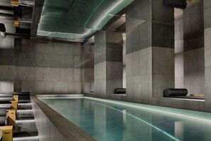 Une piscine intérieure moderne, de forme longue et étroite, entourée de hauts murs sombres et agrémentée de sièges au design minimaliste.