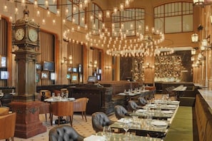 Un vaste espace dédié à la salle à manger, avec plusieurs tables, une grande horloge et des luminaires suspendus.