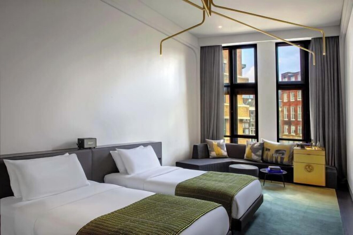 A hotel room with two beds, a sofa, a small table, and a view of buildings through large windows.