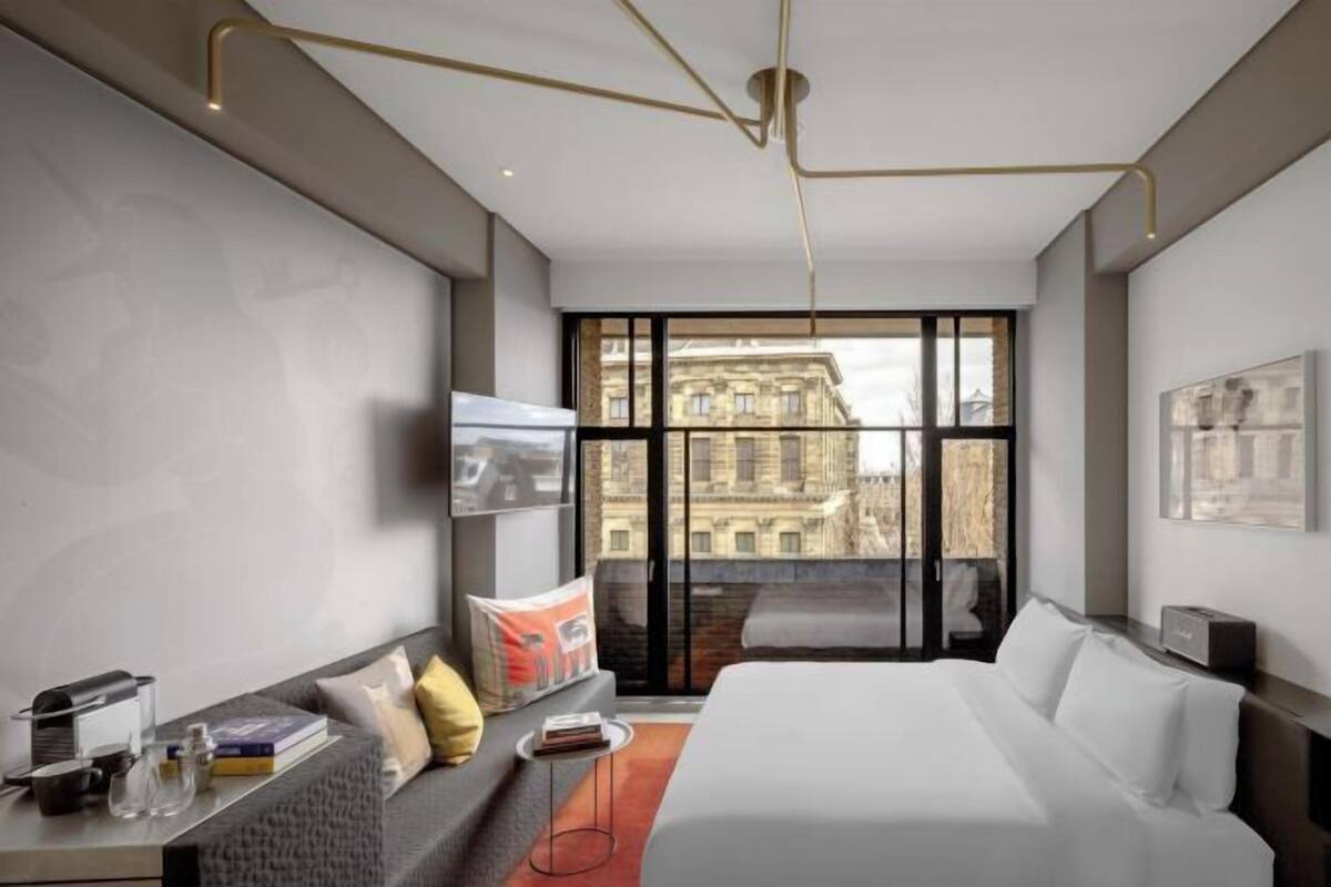 A modern hotel room with a large bed, a sofa, a TV, and a view of a cityscape through large windows.