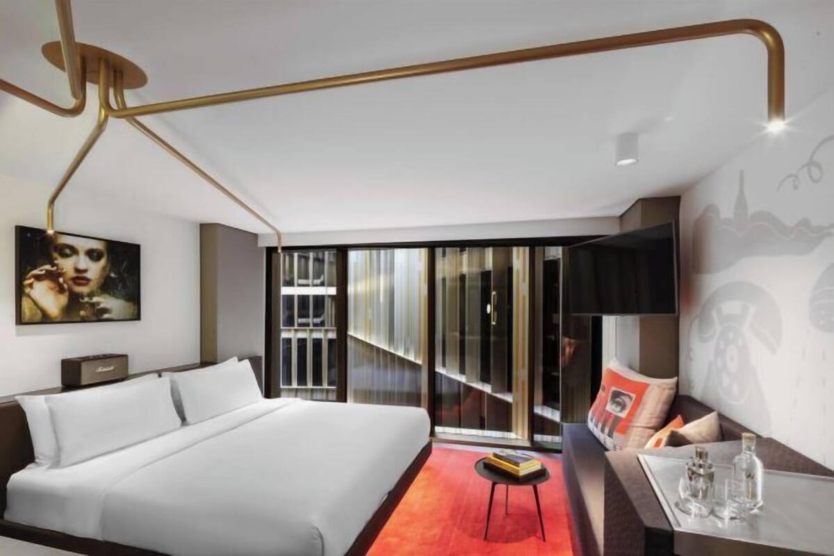 A modern hotel room with a large bed, a desk with a chair, a glass table, a painting on the wall, and a view through the glass door.