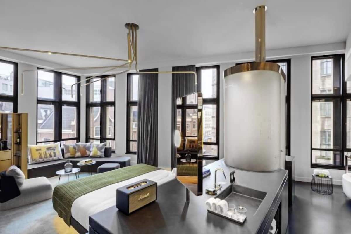 A modern hotel room with a large bed, a sitting area with a sofa and chairs, a dining table, and a large window offering a city view.