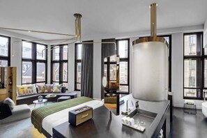 Een moderne hotelkamer met een groot bed, een zithoek met een bank en stoelen, een eettafel en een groot raam met uitzicht op de stad.