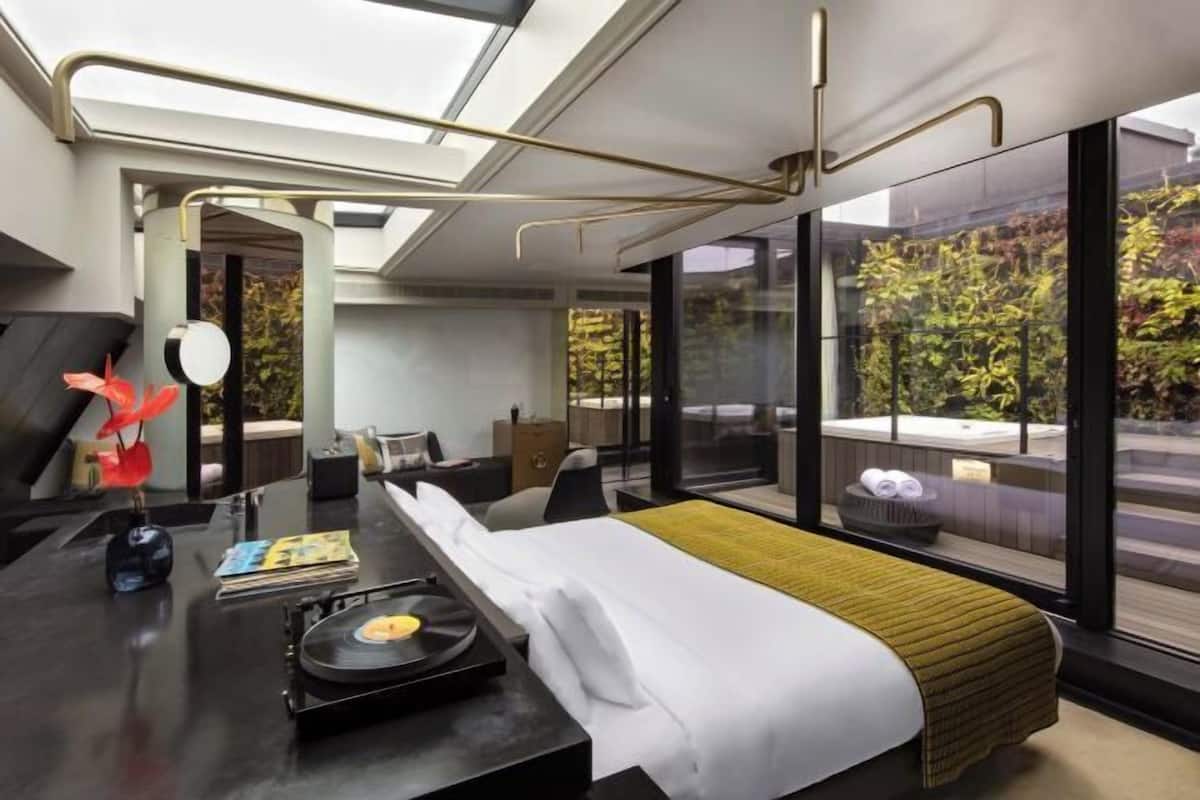 A modern hotel room with a large bed, a desk with a record player, a view of a garden through large windows, and a hot tub visible through the glass.