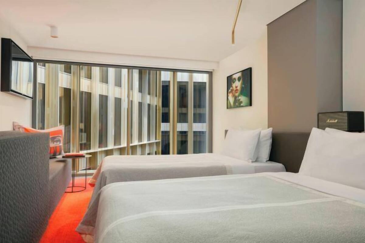 A hotel room with two beds, a sofa, a TV mounted on the wall, and a large window with blinds.