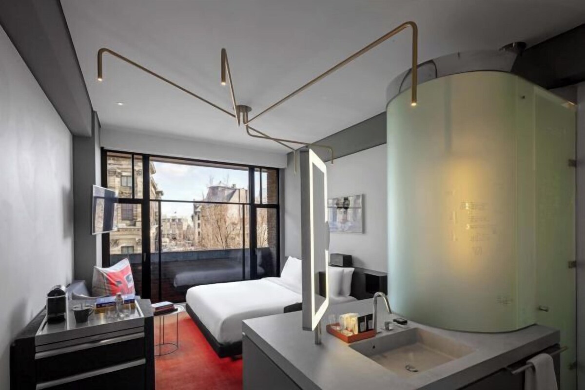 A modern hotel room with a large window, a bed, a bedside table with a mirror, and a bathroom with a glass shower enclosure.