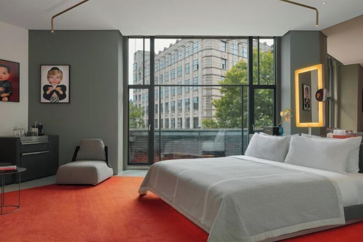 A modern hotel room with a large window offering a city view, a bed with white bedding, a desk with a chair, and a small sofa.