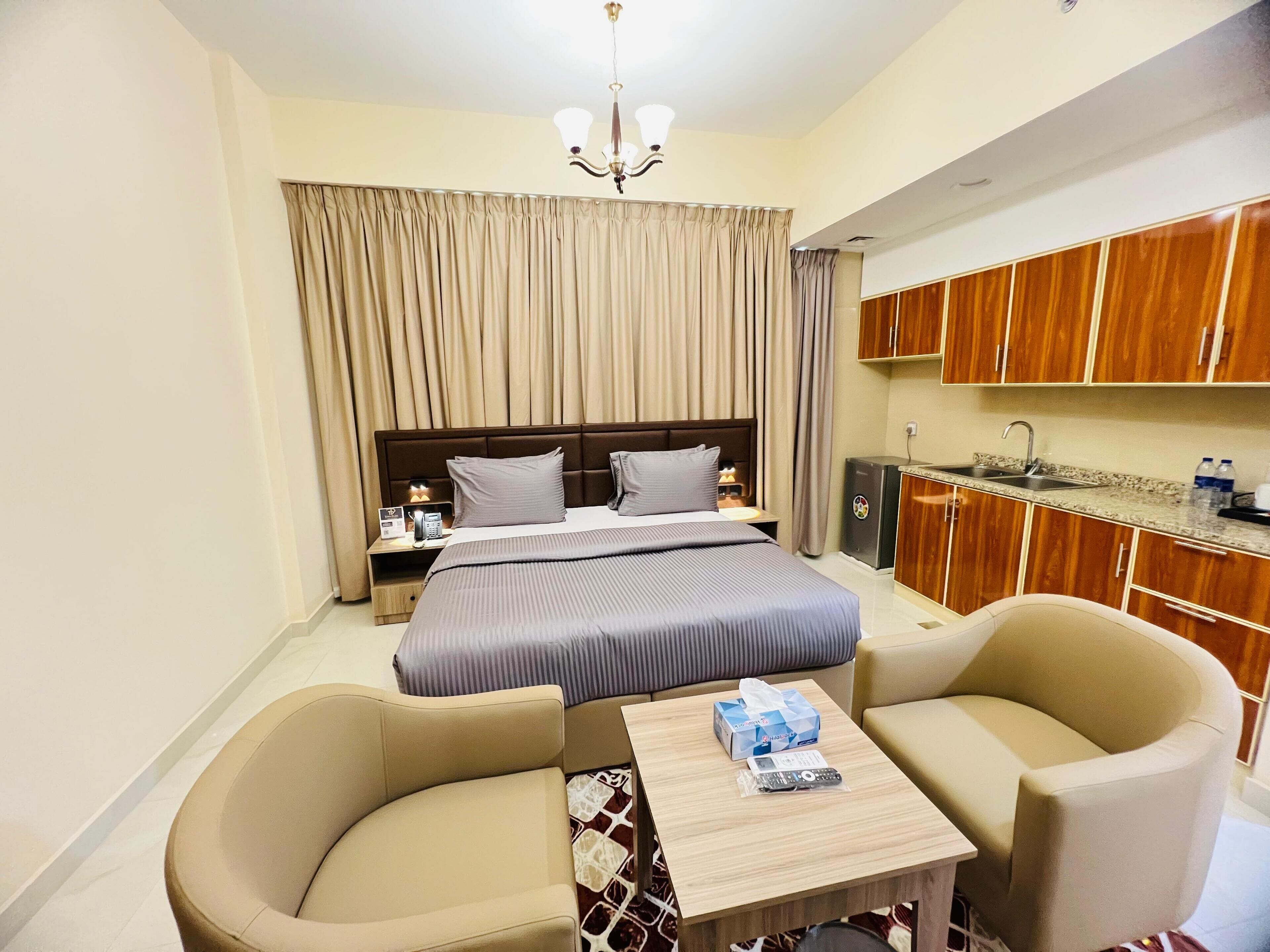 A hotel room with a bed, bedside table, lamp, curtains, a small kitchenette, and two chairs.